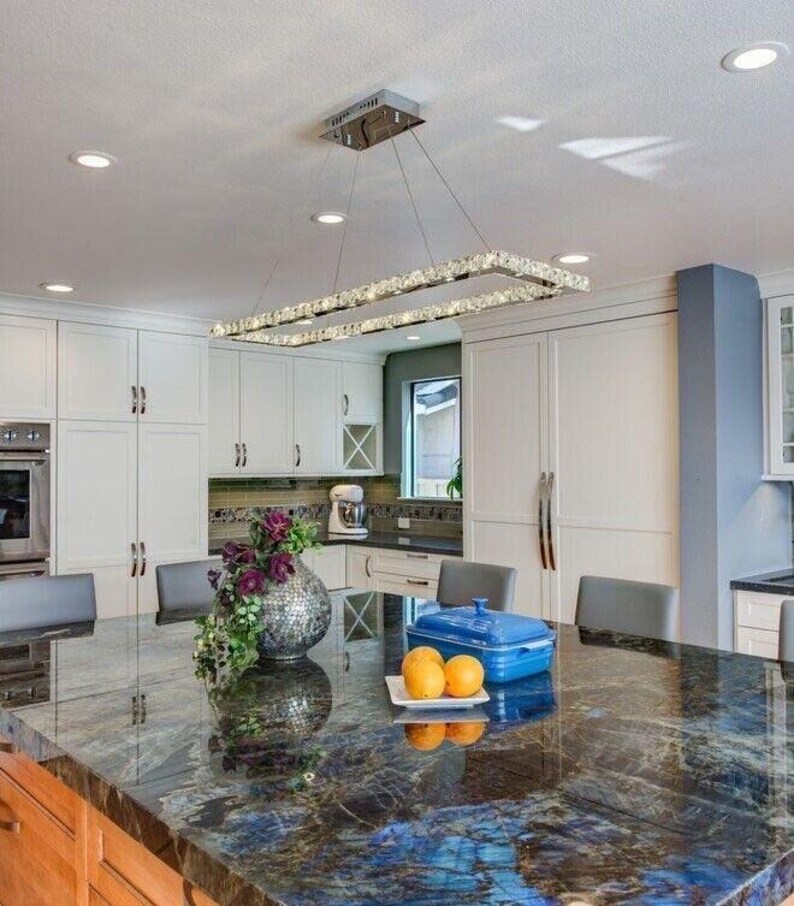 May include: A rectangular crystal chandelier hangs above a kitchen island with a dark blue and black granite countertop. On the island sits a silver textured vase with purple flowers, a blue rectangular lidded dish, and two yellow oranges.