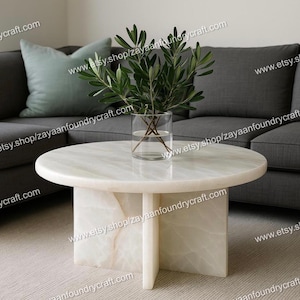 May include: A round, white marble coffee table with a pedestal base. A clear glass vase with green leafy branches sits on the table. The table is in front of a gray sofa and a light green pillow.