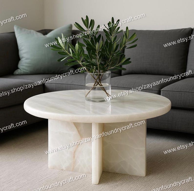 May include: A round, white marble coffee table with a pedestal base. A clear glass vase with green leafy branches sits on the table. The table is in front of a gray sofa and a light green pillow.