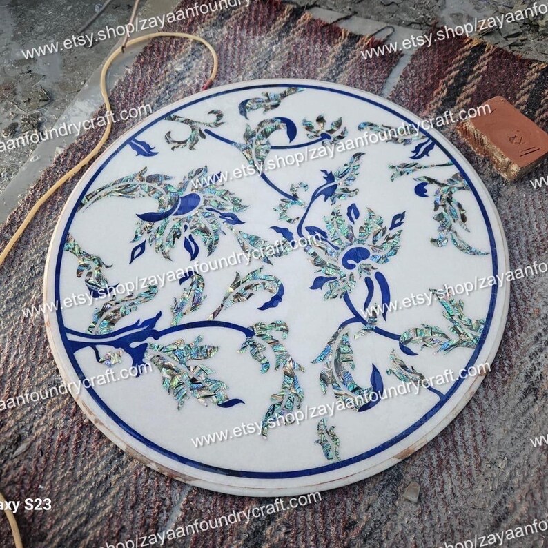 May include: A white marble table top with a blue and green floral design inlaid with mother of pearl. The design features intricate details and a delicate floral pattern.