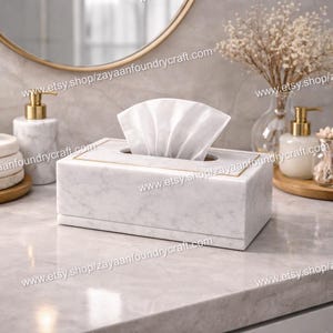 Op de afbeelding: Een rechthoekige witte marmeren tissuebox met een gouden rand, met witte tissues. De doos staat op een marmeren aanrecht, met een goudomrande spiegel en andere marmeren badkameraccessoires op de achtergrond.