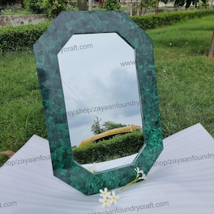 May include: An octagonal mirror with a malachite-patterned green frame. The mirror reflects a cloudy sky and a yellow vehicle. White flowers rest on the surface in front of the mirror. The text "www.etsy.shop/zayaanfoundrycraft.com" is visible.