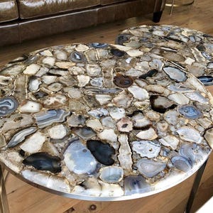 May include: Two round agate coffee tables with a gold metal base. The tabletops are made of natural agate slices in shades of brown, gray, and white.