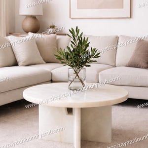 May include: A modern living room scene featuring a white marble coffee table with a cylindrical base and a glass vase holding green leafy branches. A beige sectional sofa and a framed artwork complete the neutral-toned decor.