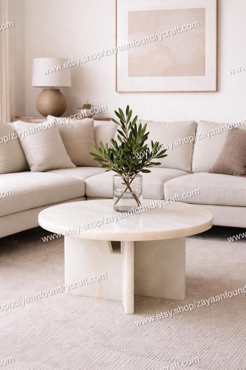 May include: A modern living room scene featuring a white marble coffee table with a cylindrical base and a glass vase holding green leafy branches. A beige sectional sofa and a framed artwork complete the neutral-toned decor.