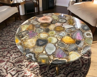 May include: A round coffee table with a unique agate stone top. The table top is made of many different colors and patterns of agate, including brown, gold, pink, and purple. The table has a gold metal base.