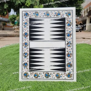 Puede incluir: Un tablero de backgammon de mármol blanco con un borde floral en azul, dorado y verde. El tablero presenta puntas triangulares negras y blancas. El texto "www.etsy.shop/zayaanfoundrycraft.com" es visible.