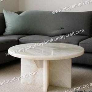 May include: A round, white marble coffee table with a pedestal base. The table is set in front of a gray sofa with a large, light green pillow. The table's dimensions are approximately 36 inches in diameter.
