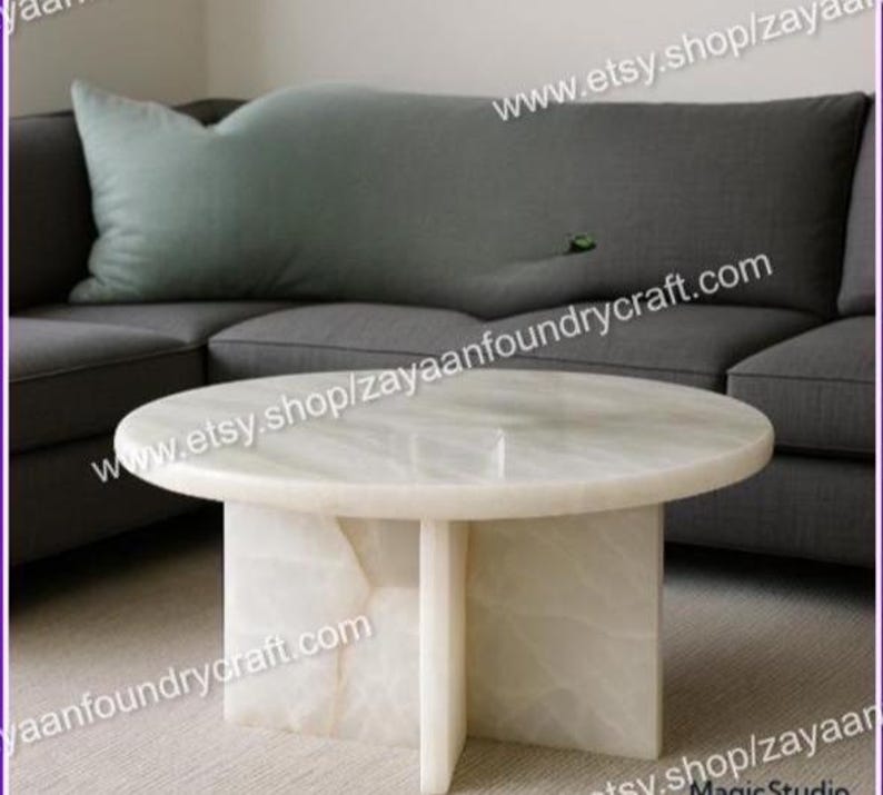 May include: A round, white marble coffee table with a pedestal base. The table is set in front of a gray sofa with a large, light green pillow. The table's dimensions are approximately 36 inches in diameter.