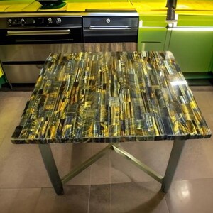 May include: A square table with a patterned top in shades of gold, blue, and black. The table has a polished surface and is supported by four silver metal legs. The table is in a kitchen setting.