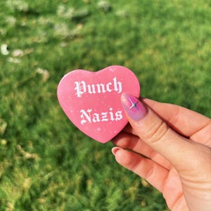 May include: A pink heart-shaped button with the words "Punch Nazis" in white gothic lettering. The button is held against a background of green grass.