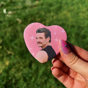 May include: A pink heart-shaped button featuring a portrait of a man with a mustache and dark hair. The background is pink with white starbursts. The button is held against a green, grassy background.