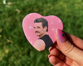May include: A pink heart-shaped button featuring a portrait of a man with a mustache and dark hair. The background is pink with white starbursts. The button is held against a green, grassy background.