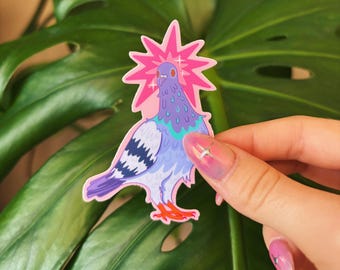 May include: A colorful sticker featuring a cartoon pigeon with a pink burst radiating from its head. The pigeon has purple, blue, and orange accents. The sticker is held in front of a large green leaf.