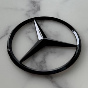 Peut inclure: Un emblème Mercedes-Benz noir brillant, représentant une étoile à trois branches dans un cercle. L'emblème est posé sur un fond de marbre blanc et gris. Les pointes de l'étoile sont nettes et réfléchissantes.
