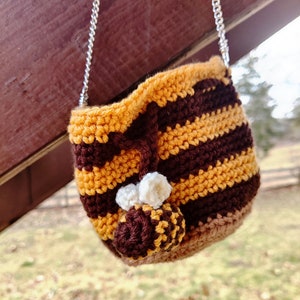 May include: A small crocheted bag with a brown and yellow striped pattern. The bag has a chain strap and a crocheted bee charm.