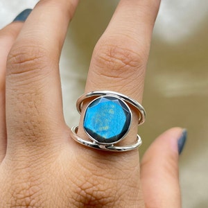 以下が含まれることがあります： 青と緑の輝きを持つ、大きな楕円形のラブラドライトの宝石をあしらったシルバーリング。リングは特徴的なダブルループバンドのデザインです。宝石が中心的な特徴で、その虹色の色合いを際立たせています。