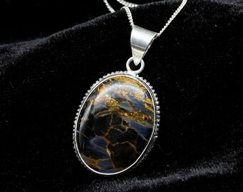 Puede incluir: Un collar con colgante ovalado que presenta una piedra preciosa pietersita pulida con patrones arremolinados en azul oscuro y marrón dorado. La piedra está engastada en un marco de plata con borde de cuentas y cuelga de una cadena fina.