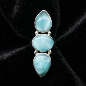 May include: A sterling silver ring featuring three polished, light blue larimar stones. The design includes a central oval stone flanked by two pear-shaped stones, all set in smooth metal bezels with small silver spheres between each setting.