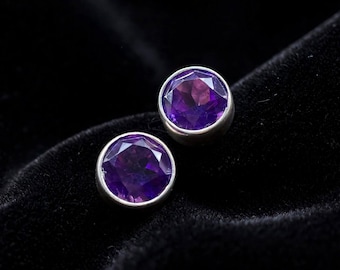 May include: A pair of round stud earrings featuring faceted purple amethyst gemstones set in smooth silver-toned metal bezels. The stones exhibit a deep violet hue with clear geometric facets that catch the light, creating a polished appearance.