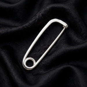 May include: A large, polished silver-toned safety pin featuring a smooth, elongated rectangular frame with rounded corners and a circular coil mechanism at the base. The metal surface is reflective, showing a bright, clean, and metallic finish.