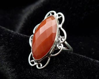 May include: A silver ring featuring a large, marquise-shaped, faceted carnelian stone. The setting is composed of intricate, swirling silver wirework that frames the central orange-red gemstone, creating a decorative, open-work metal border.