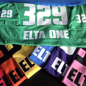 May include: A collection of short-sleeved t-shirts in various colors including green, orange, yellow, blue, purple, and pink. Each shirt features the number 329 and the text ELTA ONE printed in large, bold lettering on the front and sleeves.