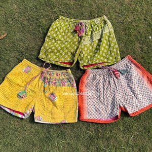 May include: Four pairs of colorful, patterned shorts are displayed on green grass. The shorts are in shades of yellow, green, and white, with decorative drawstring waists and contrasting trim. The shorts appear to be made of a lightweight fabric.