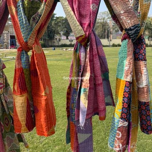 May include: Colorful, patchwork scarves hanging outdoors. The scarves are made of various fabrics in shades of orange, pink, green, and blue. Each scarf is knotted, showcasing the different patterns and textures. The text "Sarvayacreation" is visible on the image.