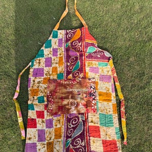 Op de afbeelding: Een kleurrijke patchwork schort met vierkante patronen in paars, oranje, rood en groen. De schort heeft een zak met de tekst "Sarvayacreation". Het is gemaakt van stof en heeft een nekband en strikbanden.