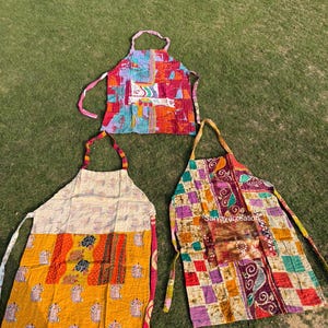 Può includere: Tre grembiuli patchwork colorati con vari motivi e disegni sono esposti su erba verde. I grembiuli presentano un mix di colori vivaci, tra cui rosso, giallo, blu e viola, con motivi unici. Ogni grembiule ha un anello per il collo e lacci in vita.