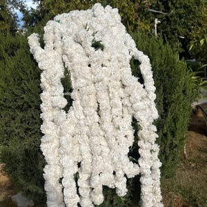 May include: A long, cascading garland of white, fluffy flowers. The garland is draped over green foliage, with the flowers appearing to be made of a soft, textured material. The overall effect is delicate and decorative, suitable for events or home decoration.