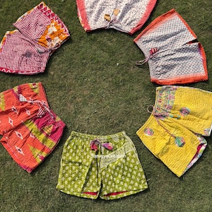 May include: A collection of colorful, patterned shorts arranged on green grass. The shorts feature various designs, including floral and geometric patterns, in shades of red, yellow, green, and white. Some shorts have drawstring closures.