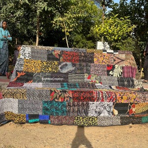May include: A patchwork bench cover with a variety of colorful fabric squares. The cover features floral and geometric patterns in shades of red, black, yellow, and blue. The bench is outdoors, with trees and a person in the background.