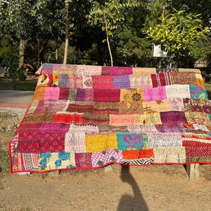 May include: A colorful patchwork quilt with a red border is draped over a bench. The quilt features a variety of fabrics in shades of pink, red, yellow, and blue. The quilt is made of many different squares of fabric.