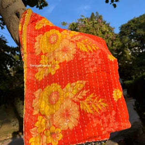 Puede incluir: Un edredón floral rojo vibrante con grandes flores amarillas y costuras blancas. El edredón está drapeado sobre una rama de árbol, mostrando su intrincado diseño y textura. Los bordes están adornados con un borde amarillo.