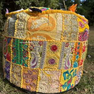Op de afbeelding: Een ronde, gele, veelkleurige, geborduurde poef met een patchwork-ontwerp. De poef is gemaakt van samen genaaide stofresten en versierd met kralen, pailletten en borduurwerk. De poef staat op een bed van groen gras.