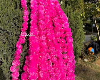 May include: Bright pink floral garland draped over a green bush. The garland is densely packed with small, round flowers, creating a vibrant and eye-catching display. The image is taken outdoors, with a glimpse of a park bench in the background.