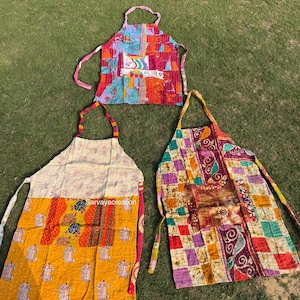 Op de afbeelding: Drie kleurrijke patchwork schorten met nek- en taillebanden worden getoond op groen gras. De schorten hebben een verscheidenheid aan patronen en kleuren, waaronder bloemen-, geometrische en abstracte ontwerpen. De tekst "Sarvayacreation" is zichtbaar op een schort.