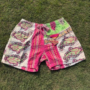 May include: Paisley print shorts with a drawstring waist. The shorts feature a mix of colors, including pink, green, and white. The fabric has a quilted texture. The shorts are laying on a bed of green grass.