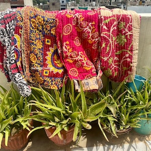 Può includere: Una collezione di trapunte colorate e decorate appese all'aperto. Le trapunte presentano tonalità vibranti di rosso, giallo, blu e verde con disegni floreali e geometrici. Piante di aloe vera in vaso in primo piano.