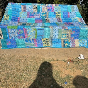 Puede incluir: Una colcha de retazos con una variedad de telas azules, rosas y verdes. La colcha está hecha de muchas piezas diferentes de tela cosidas juntas para crear un diseño único. La colcha está extendida en el suelo frente a una zona de hierba.