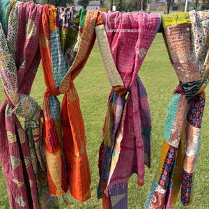Puede incluir: Una colección de coloridas bufandas de patchwork colgadas al aire libre. Las bufandas presentan una variedad de patrones y colores, incluyendo rosa, naranja, verde y azul. La imagen está tomada en un día soleado con hierba verde en el fondo.