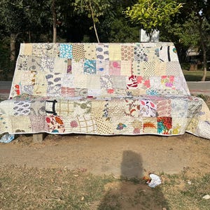 May include: A patchwork quilt made of various fabrics in shades of white, blue, pink, red, and brown. The quilt is laid out on a bench in a park setting.