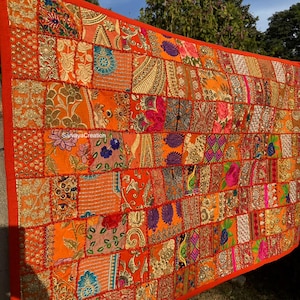 Op de afbeelding: Een grote, rechthoekige patchwork wandhanger met een feloranje achtergrond. De patchwork is gemaakt van verschillende stoffen in tinten oranje, rood, roze, paars en goud. De stof is versierd met ingewikkelde borduursels en pailletten. De wandhanger heeft een rode rand.
