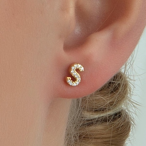 May include: Gold stud earrings featuring the letter 'S' encrusted with small clear cubic zirconia stones.