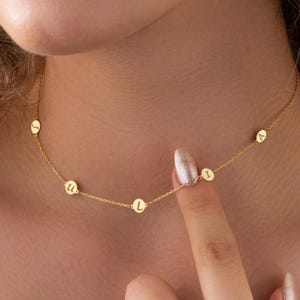 May include: A delicate gold-colored necklace featuring small, circular pendants with engraved letters. The necklace is worn around the neck, with the letters 'J', 'U', 'L', 'I', and 'A' visible. The chain is thin and the letters are in a simple font.
