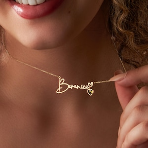 May include: A gold necklace with a delicate chain and a pendant featuring the name "Berenice" written in cursive script. A small heart charm with a green gemstone hangs from the end of the name.