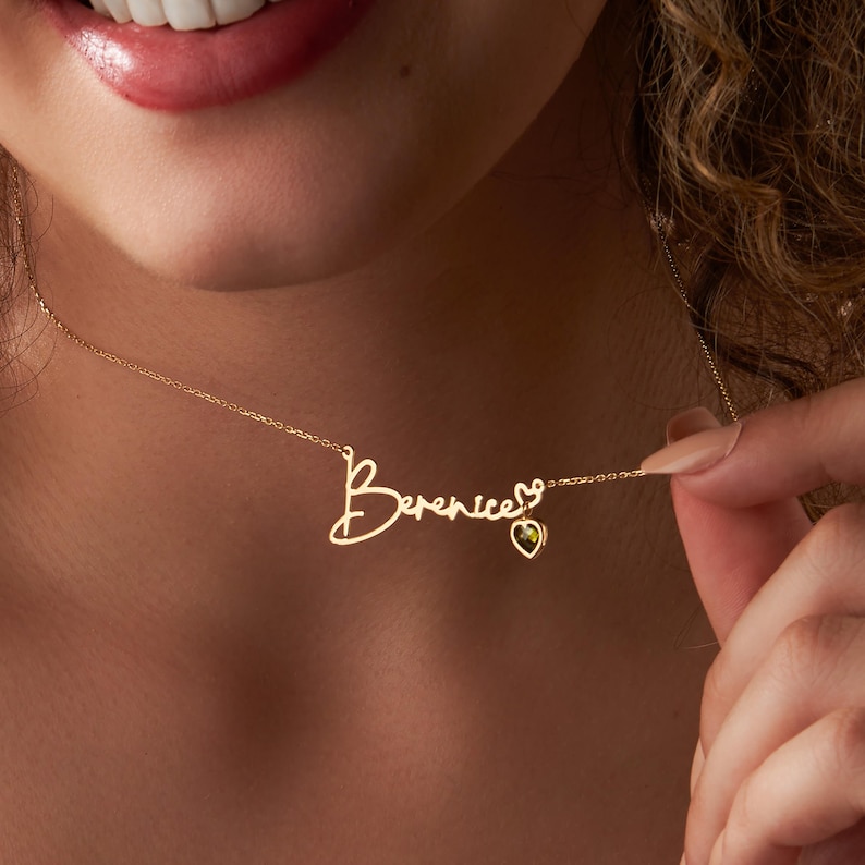 May include: A gold necklace with a delicate chain and a pendant featuring the name "Berenice" written in cursive script. A small heart charm with a green gemstone hangs from the end of the name.