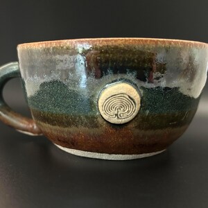 May include: A brown and green ceramic mug with a white glaze and a small, white, circular design with a labyrinth pattern in the centre.
